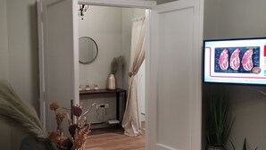 Um corredor com piso de madeira, espelho na parede, uma pequena prateleira com objetos decorativos e uma televisão exibindo comida.