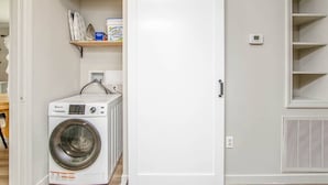 Ein Waschraum mit einer Waschmaschine mit Frontlader, einem Regal für Waschmittel und einem Thermostat an der Wand.