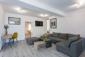 A modern living room with a grey sectional sofa, a wooden coffee table, and a yellow chair. There is a dining table with chairs and a TV mounted on the wall.