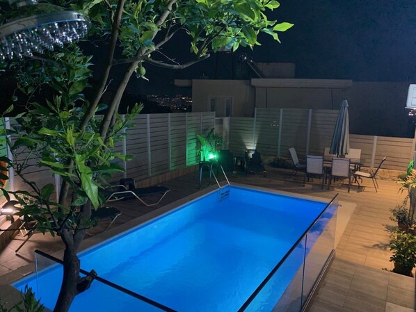 A swimming pool with a wooden deck, lounge chairs, and a green-lit wall.