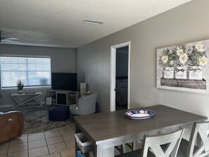 A living room with a TV, a dining table, and a painting on the wall. - Teak House-Relaxing under the Sycamore Trees/Pet Friendly (Ocala)