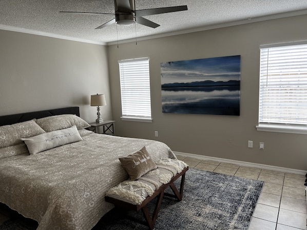 A bedroom with a bed, a bench, a ceiling fan, and a large landscape painting. - Teak House-Relaxing under the Sycamore Trees/Pet Friendly (Ocala)
