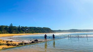 A beach with clear skies, rocky shoreline, and a distant forested hill. - Feel The Aroha-Love At This Beautiful Beach Haven (MACMASTERS BEACH)