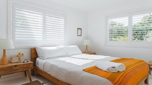 A bedroom with a wooden bed, white bedding, two bedside tables with lamps, a small vase with flowers, and a window with blinds. - Feel The Aroha-Love At This Beautiful Beach Haven (MACMASTERS BEACH)