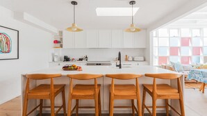 A modern kitchen with white cabinetry, a central island with bar stools, and pendant lighting. - Feel The Aroha-Love At This Beautiful Beach Haven (MACMASTERS BEACH)