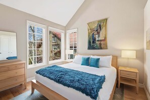 Een slaapkamer met een bed, een dressoir, een nachtkastje, een lamp en een raam met uitzicht op een huis.