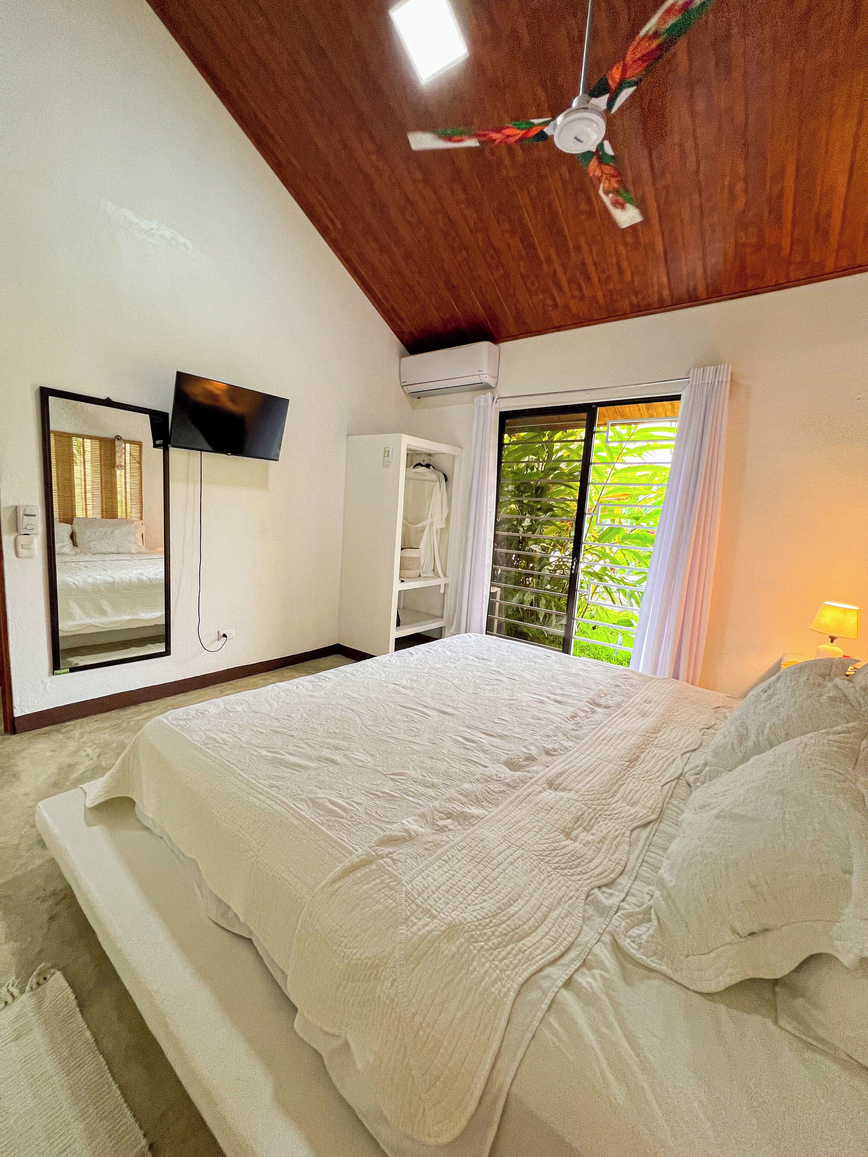 A bedroom with a bed, a ceiling fan, a TV mounted on the wall, and a window with curtains.