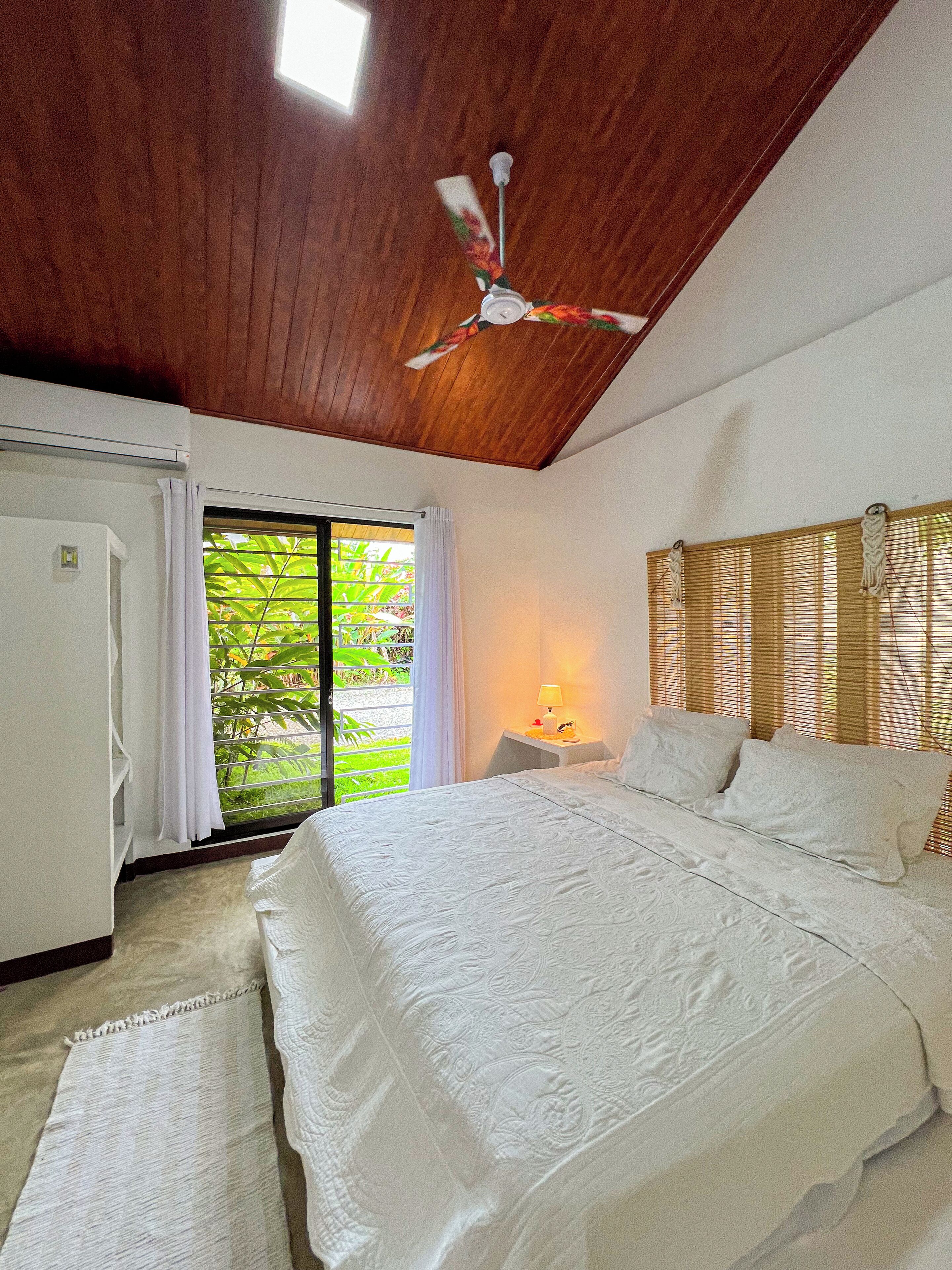 Une chambre à coucher comprenant un lit, un ventilateur de plafond, une fenêtre avec des stores et une table de chevet avec une lampe.