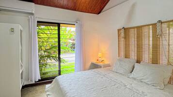 Une chambre à coucher comprenant un lit, un ventilateur de plafond, une fenêtre avec des stores et une table de chevet avec une lampe.