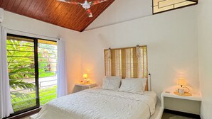 Un dormitorio con techo de madera, una cama con ropa de cama blanca, dos mesitas de noche con lámparas y vistas a la vegetación a través de una puerta corredera.
