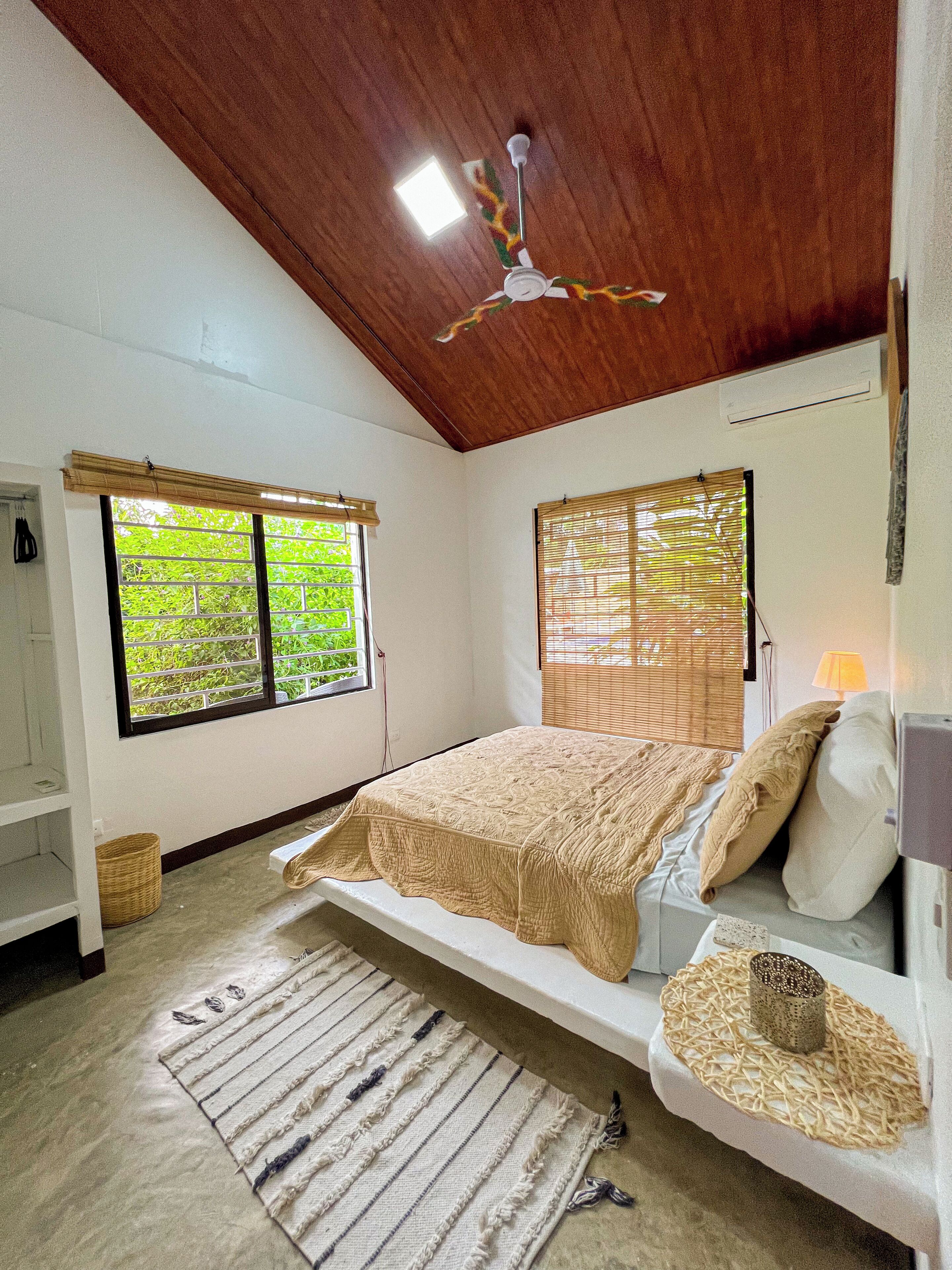 A bedroom with a bed, a ceiling fan, two windows with blinds, a bedside table with a lamp, and a woven mat.