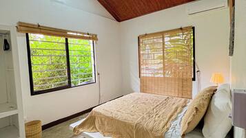 A bedroom with a bed, a ceiling fan, two windows with blinds, a bedside table with a lamp, and a woven mat.