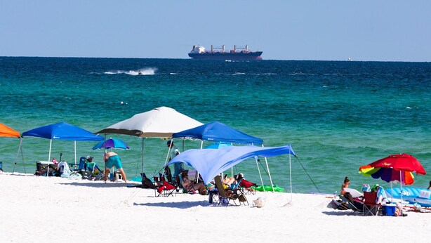 A beach with several tents and umbrellas, a large ship in the background, and people relaxing on the sand.