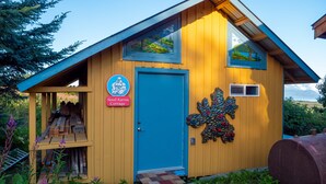 Una pequeña cabaña de madera con puerta azul, un letrero que dice 'Good Karma Cottage' y un diseño decorativo de hojas en la pared.