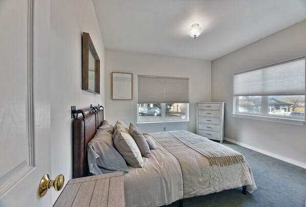 Une chambre à coucher comprenant un lit, une commode, une fenêtre avec des stores et une porte dotée d’une poignée en laiton.