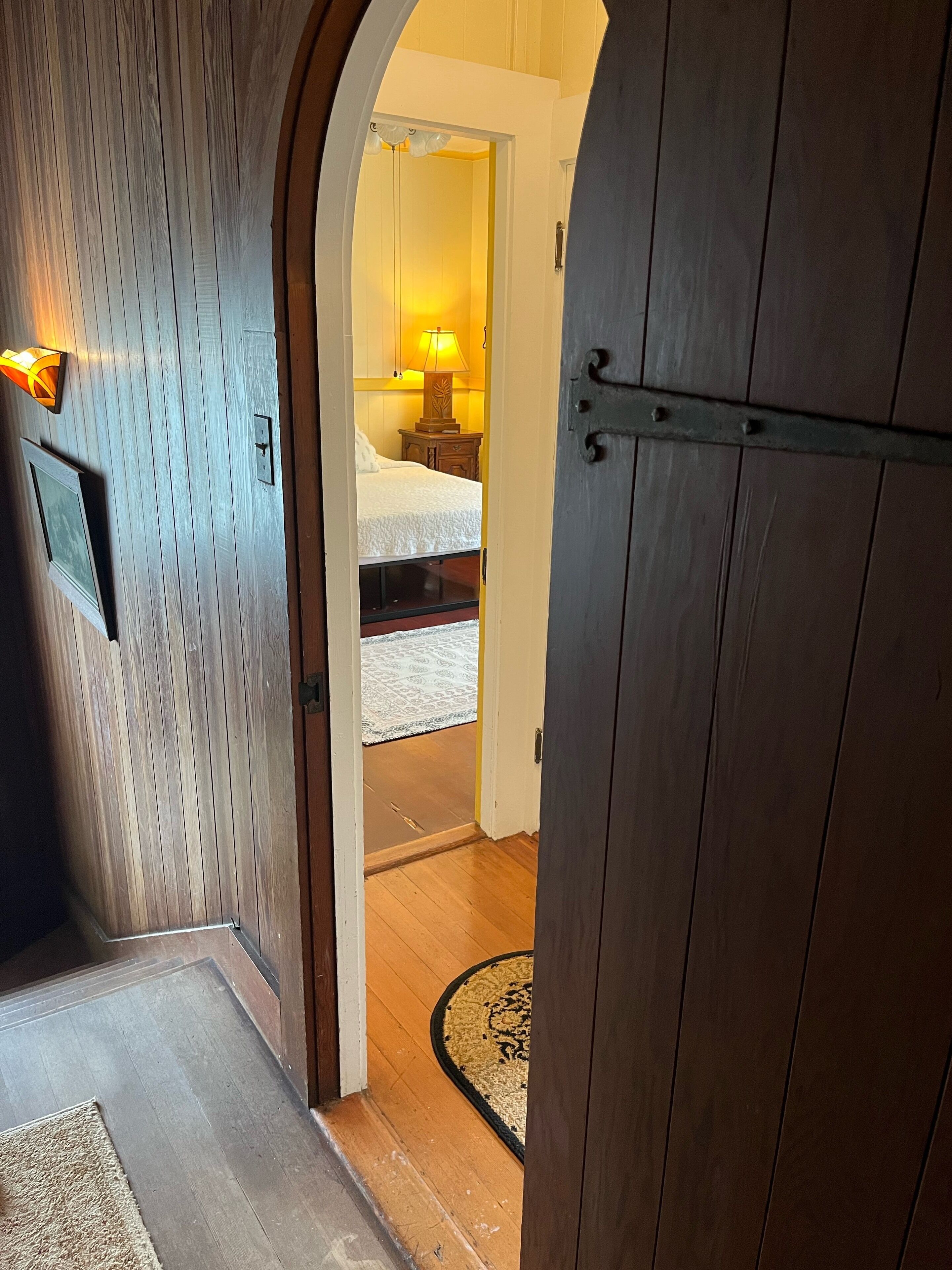 A wooden interior hallway leading to a bedroom with a bed, bedside lamp, and a nightstand.