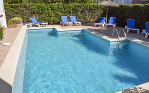 Uma piscina retangular com água azul cristalina, rodeada por espreguiçadeiras brancas e um guarda-sol de palha.