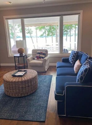A living room with a blue sofa, a round ottoman, a chair, and a large window with a view of trees and a deck.