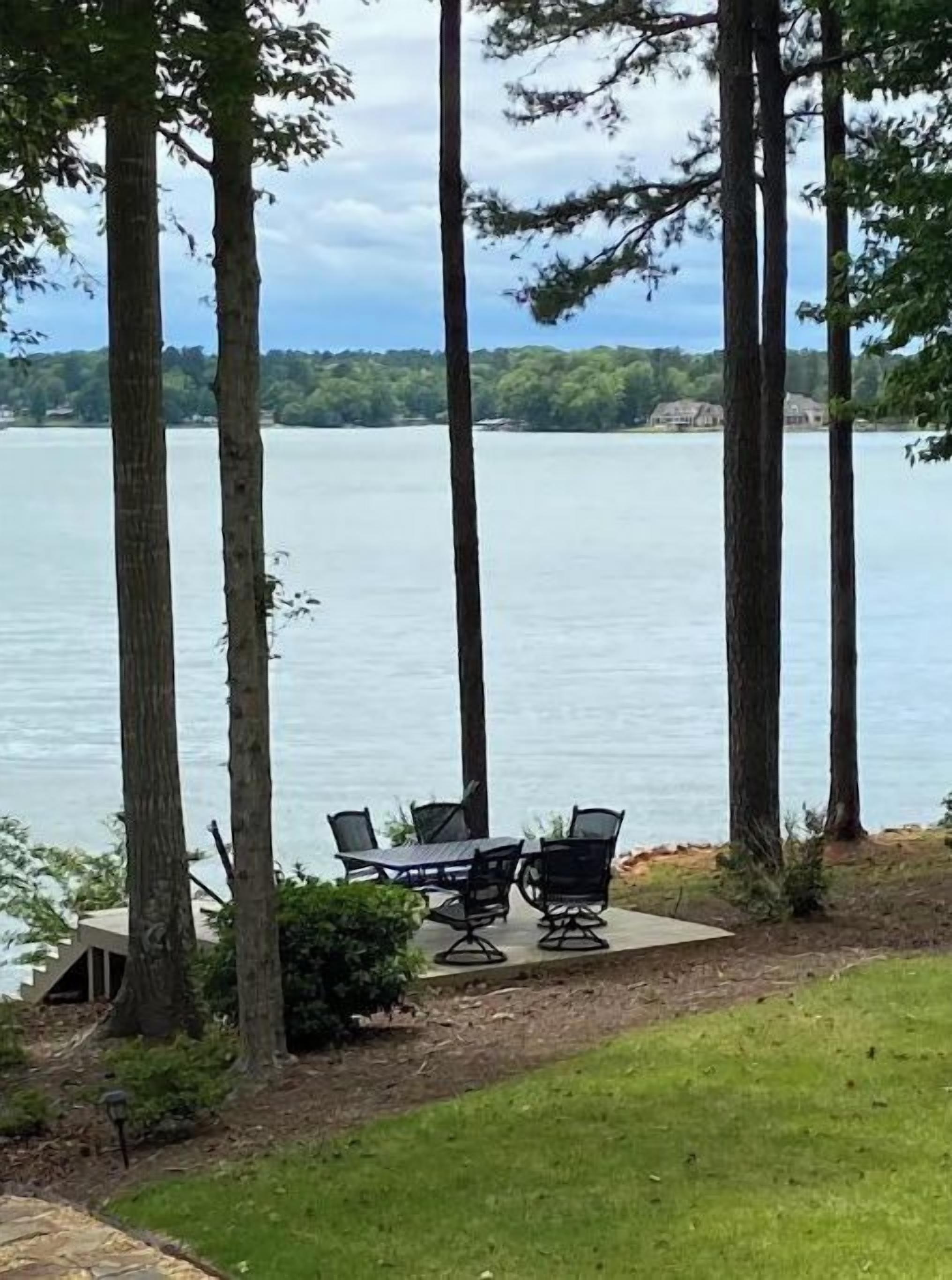 Une vue sur le lac avec un espace repas comprenant une table et des chaises, le tout entouré de grands arbres et bordé d’une pelouse.