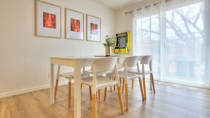 A dining area with a wooden table and white chairs, framed pictures on the wall, and a window with a view of trees.