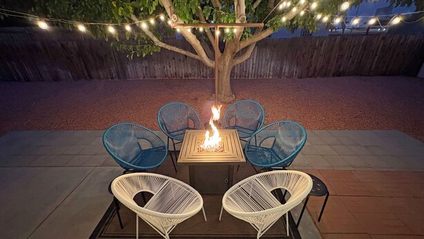 A patio with a fire pit table and four chairs, surrounded by a wooden fence and string lights.