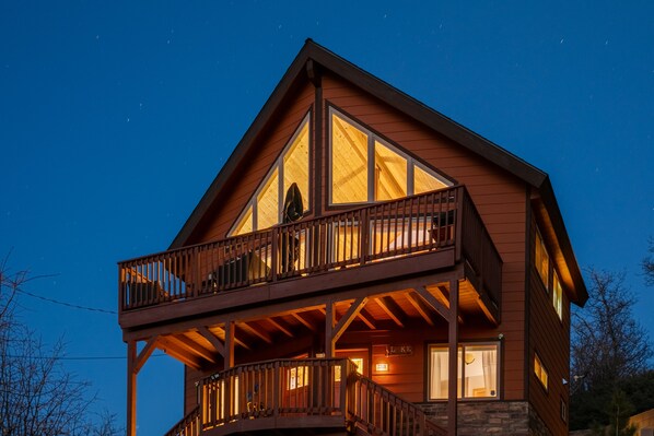 A two-story wooden cabin with a balcony, large windows, and a stone foundation.