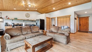 A spacious living area with a wooden ceiling, a large sofa set, a coffee table, and a kitchen in the background. - Avalanche Cabin @ Explorer's Basecamp (Kalispell)