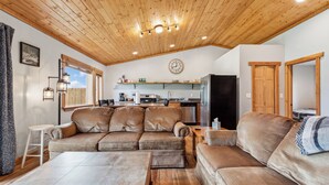 A cozy living room with a wooden ceiling, a large beige sectional sofa, a wooden coffee table, and a kitchen in the background. - Avalanche Cabin @ Explorer's Basecamp (Kalispell)