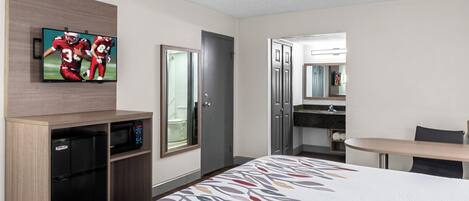 A hotel room with a bed, a TV mounted on the wall, a microwave, a small fridge, a bathroom with a mirror, and a desk with a chair.
