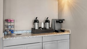 Una kitchenette con cafetera, dos termos, un portavasos y un estante con objetos.