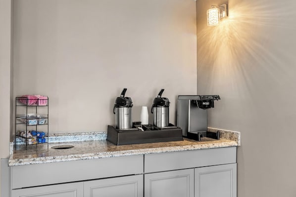 A kitchenette with a coffee maker, two thermos flasks, a cup holder, and a small shelf with items.