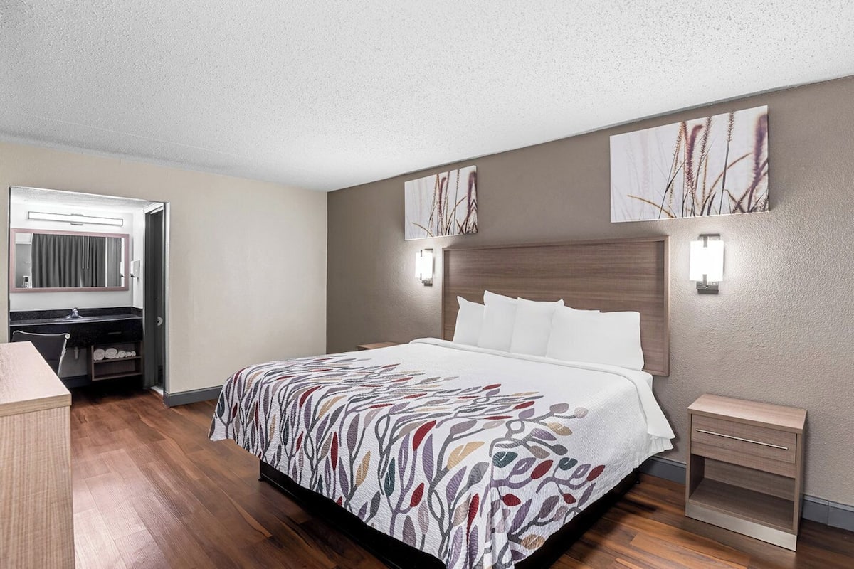 A hotel room with a large bed, a desk with a chair, a small bedside table, and two wall-mounted lights.