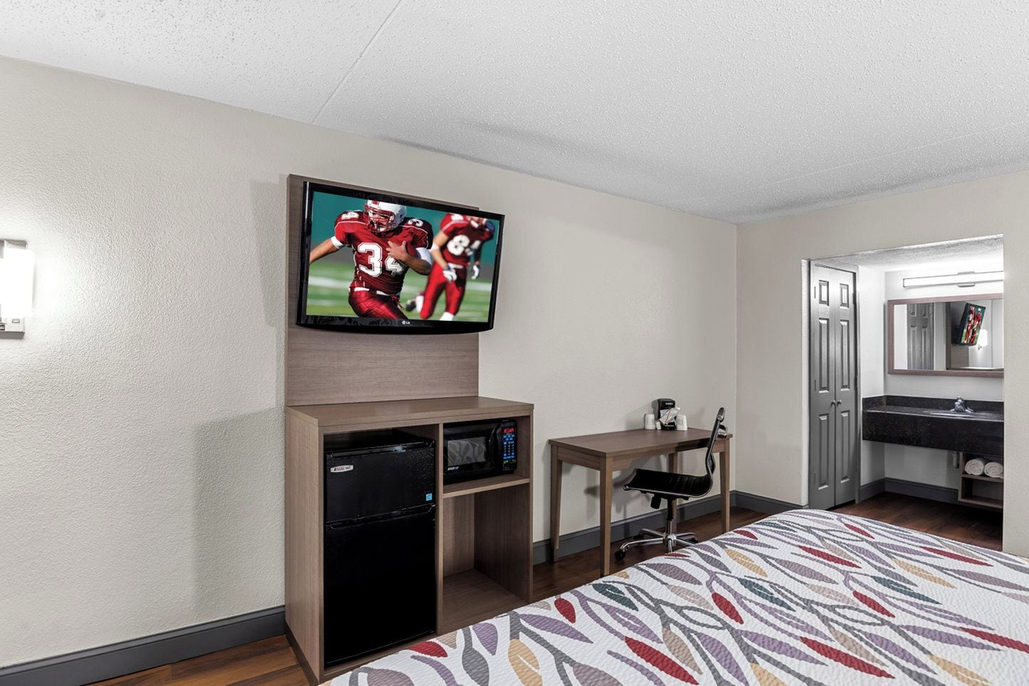 Hotelkamer met een flatscreen televisie aan de muur, een klein tafeltje met een stoel, een bureau met een stoel, een bed met een beddegoed met een patroon, en een badkamer met een spiegel en wastafel.
