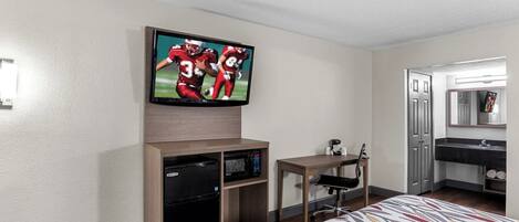 A hotel room with a flat-screen TV mounted on the wall, a small table with a chair, a desk with a chair, a bed with a patterned bedspread, and a bathroom with a mirror and sink.