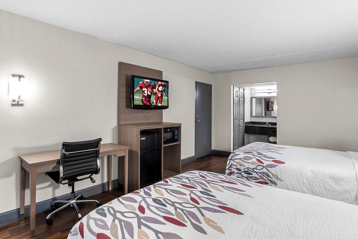 A hotel room with two beds, a desk, a chair, a television, a microwave, and a bathroom with a mirror.