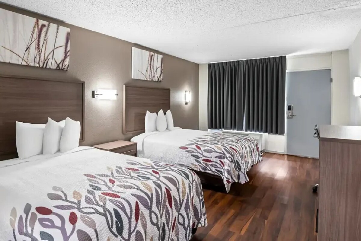 A hotel room with two beds, a wooden headboard, a large window with curtains, and a desk with a chair.