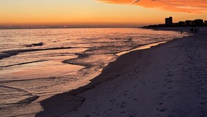 En strand med en solnedgångshimel, ett lugnt hav och byggnader i fjärran.
