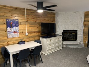 Una habitación con una mesa y sillas blancas, un televisor negro, una chimenea de piedra y un cartel que anuncia clases y alquiler de esquí.