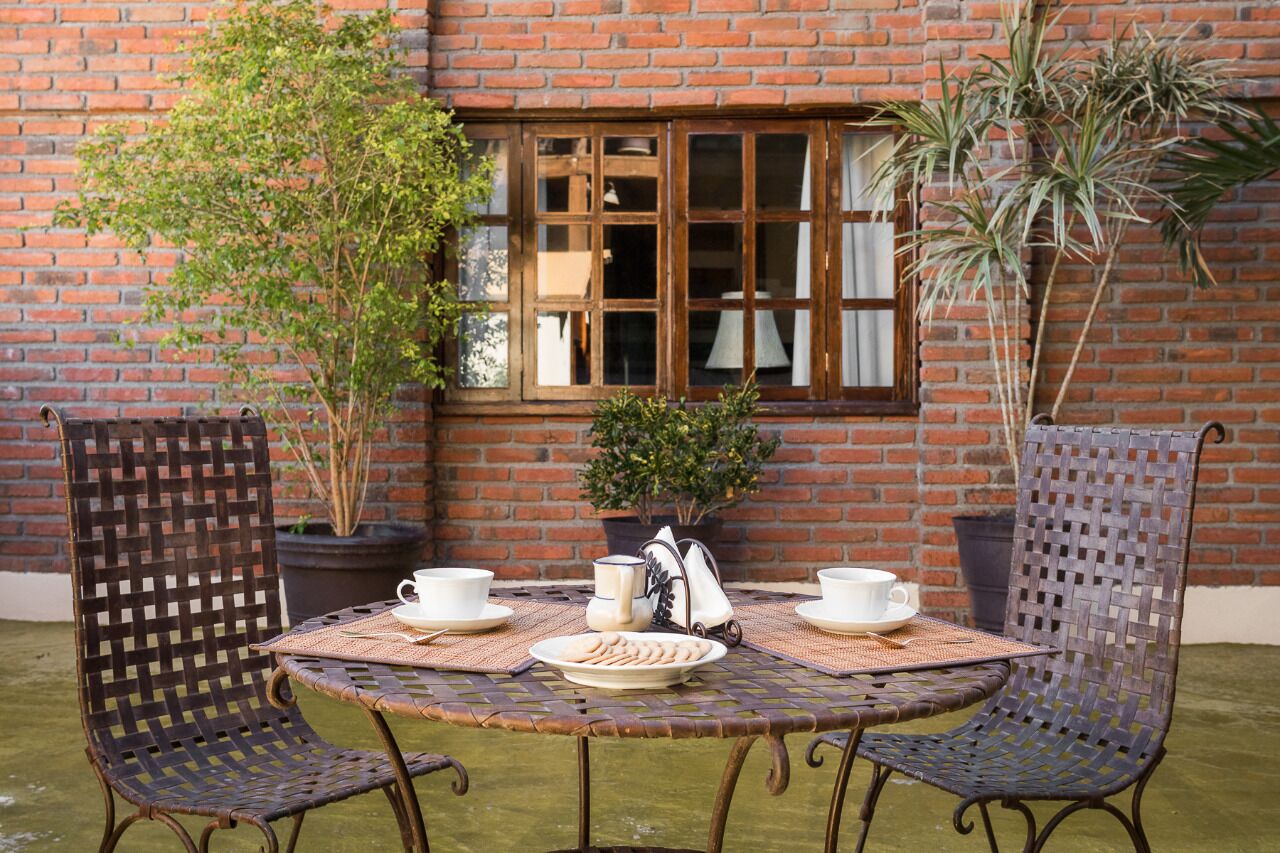 Une table ronde en métal, recouverte d’une nappe tissée, est entourée de deux chaises en osier. Sur la table, on trouve deux tasses et soucoupes blanches, un pichet blanc et une assiette remplie de biscuits.
