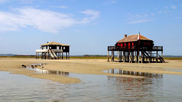 Zwei hölzerne Gebäude mit Balkonen auf Stelzen über einem Sandstrand.