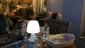 Ein Terrassenbereich im Freien mit einem Esstisch, Wein gläsern, Flaschen und einer Schüssel. Im Hintergrund befinden sich Kübelpflanzen und ein Holzzaun.