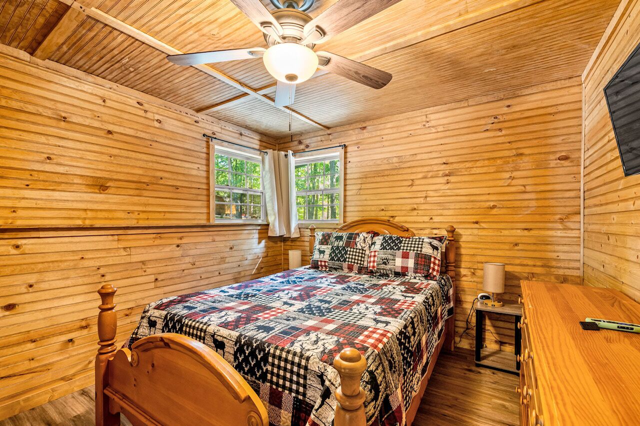 Un dormitorio de cabaña de madera con una cama, un ventilador de techo y dos ventanas.