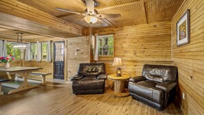 A wooden cabin interior with a ceiling fan, a dining table with benches, two leather chairs, a small round table with a lamp, and a framed picture on the wall.