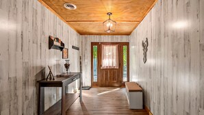 A hallway with wooden flooring, a wooden ceiling, a bench, a table with a lamp, and a door with a window.