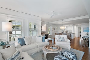 A spacious living room with a white sofa set, a round coffee table, and a dining area with a kitchen in the background.