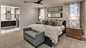 A bedroom with a bed, a bedside table, a chair, a ceiling fan, and a bathroom with a sink and mirror.