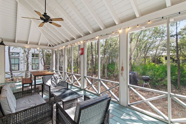 A covered outdoor area with wicker furniture, a ceiling fan, and large windows overlooking a garden.