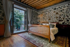Quarto com parede de pedra, teto de madeira, cama com colchão e almofadas brancas, e vista através de uma porta de vidro.