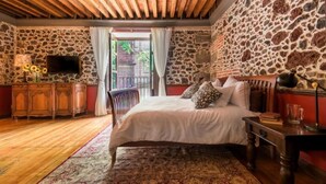 A bedroom with a stone wall, a wooden ceiling, a bed with white bedding, a nightstand, a TV, and a view of the outside. - Casa Sauto (Mexico City)