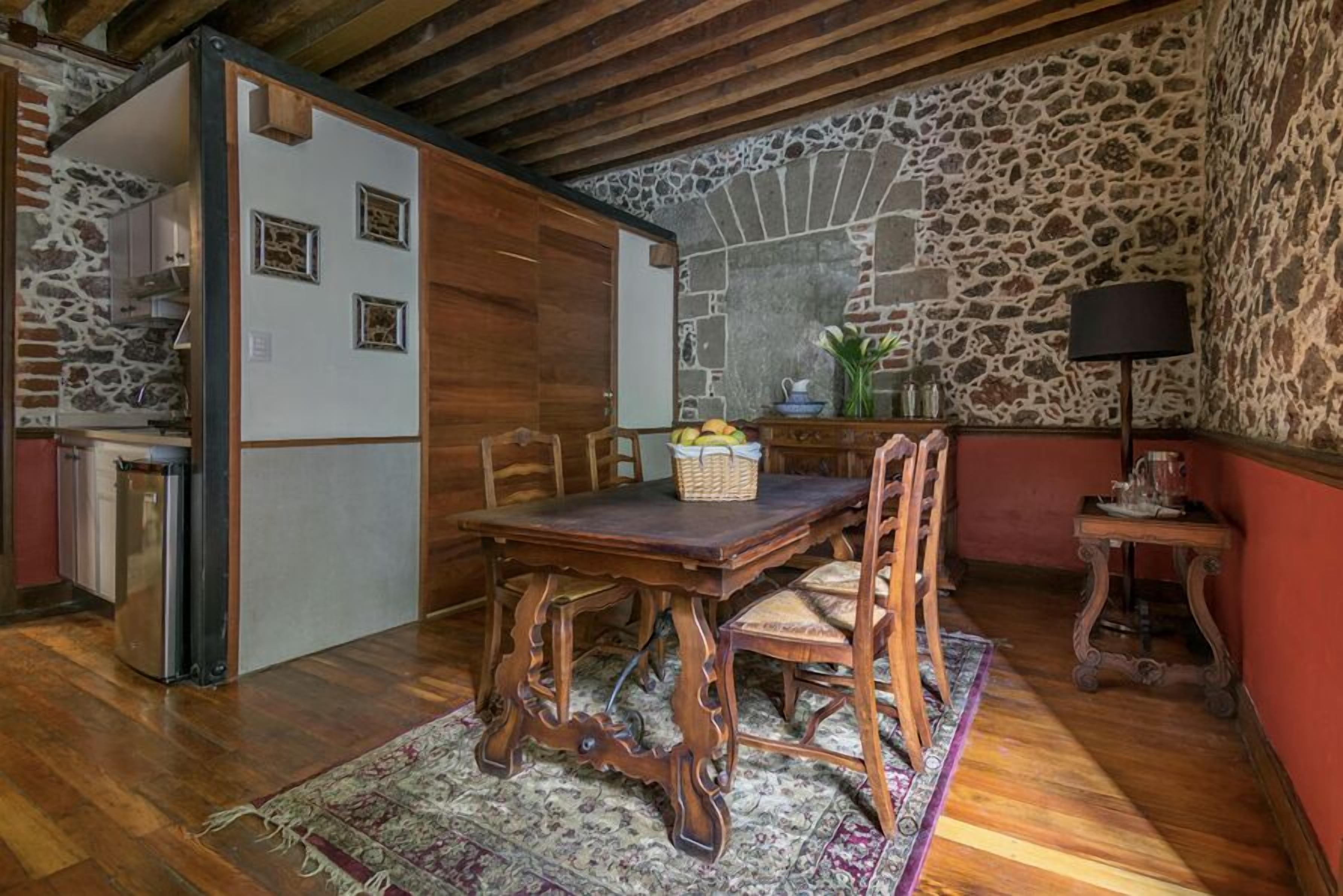 A rustic dining area with a wooden table and chairs, a stone wall, and a small kitchen area with a refrigerator and a sink.